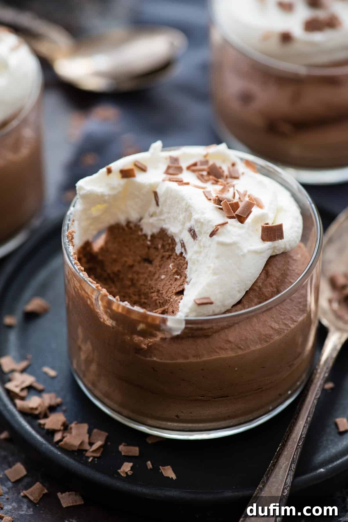 A glass dessert bowl containing chocolate mousse with a scoop removed, showcasing its creamy, airy texture. The mousse is garnished with whipped cream and chocolate shavings on a dark plate beside a metal spoon.