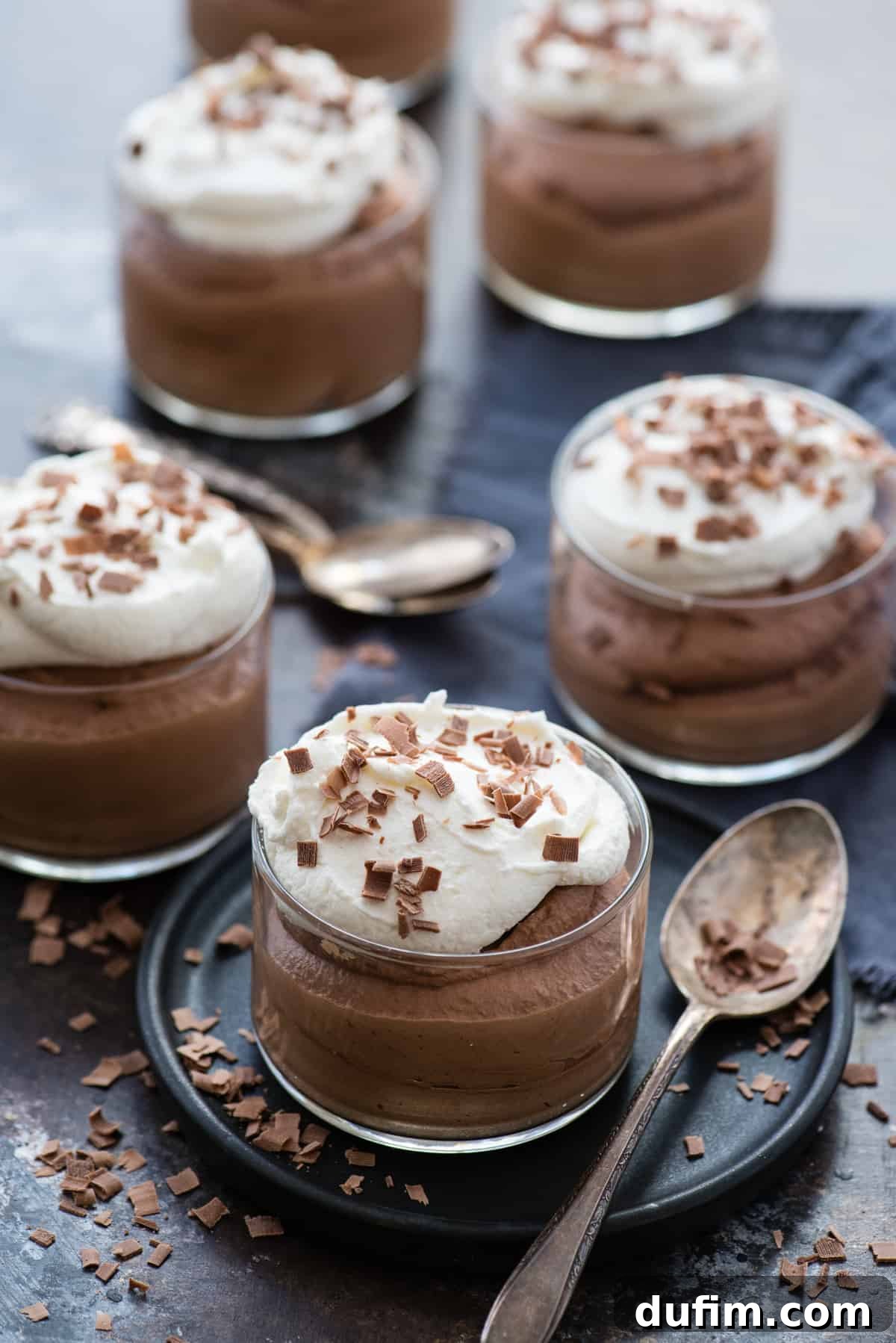 Six individual glass dessert bowls filled with creamy chocolate mousse, each topped with a dollop of whipped cream and chocolate shavings, arranged on a dark serving platter. A metal spoon rests nearby.