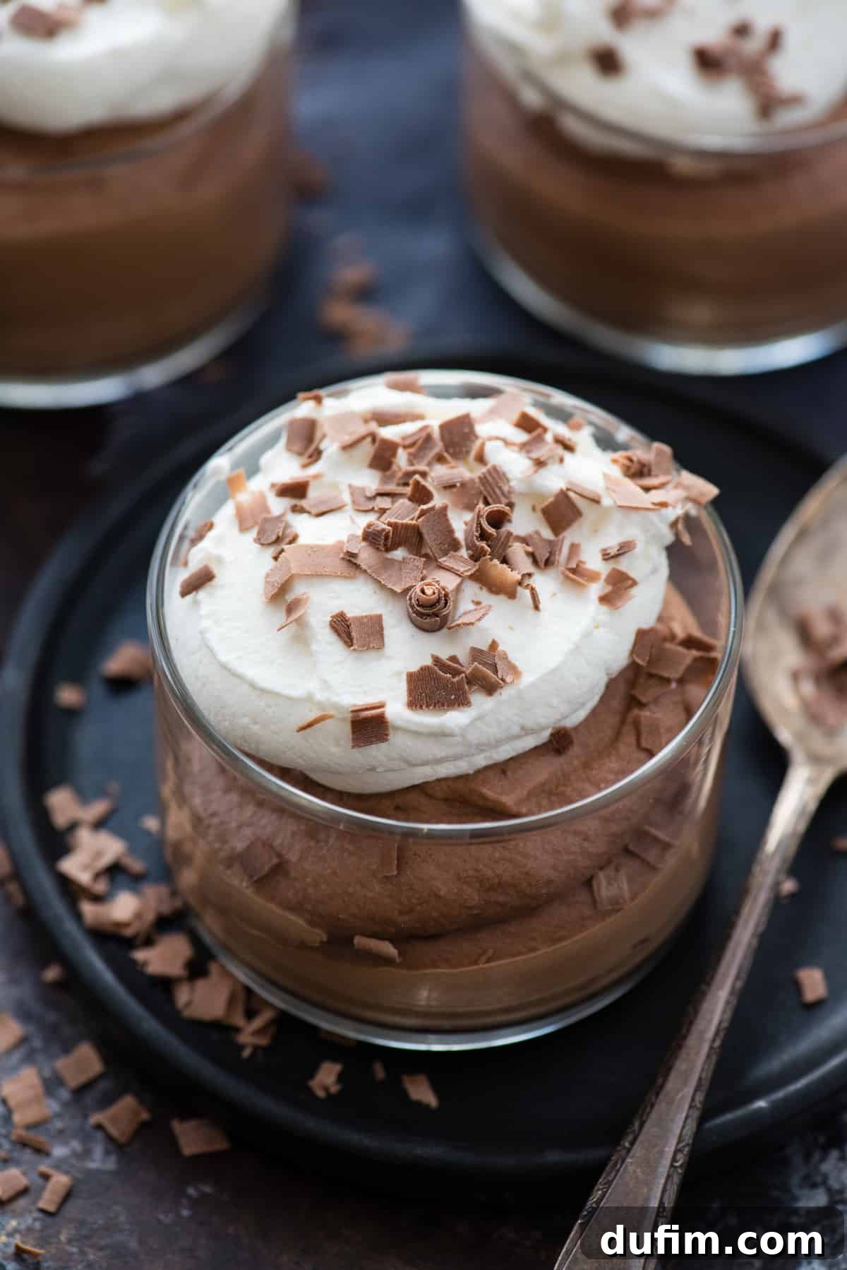 Close-up shot of a single serving of chocolate mousse in a glass bowl, garnished with whipped cream and chocolate curls, on a dark plate with a spoon. The focus highlights the smooth, rich texture of the mousse.