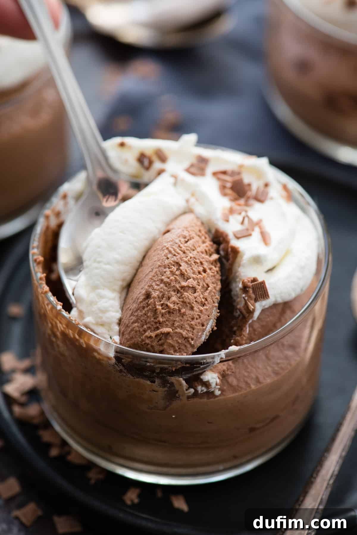 A scoop has been taken from a glass bowl of chocolate mousse, revealing its smooth and creamy interior. The mousse is topped with whipped cream and chocolate shavings, resting on a dark plate with a metal spoon, emphasizing its rich texture.