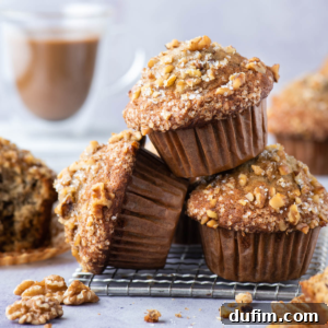 Three streusel-topped Banana Nut Muffins stacked on a cooling rack, with walnuts scattered around and a blurred cup of coffee in the background.