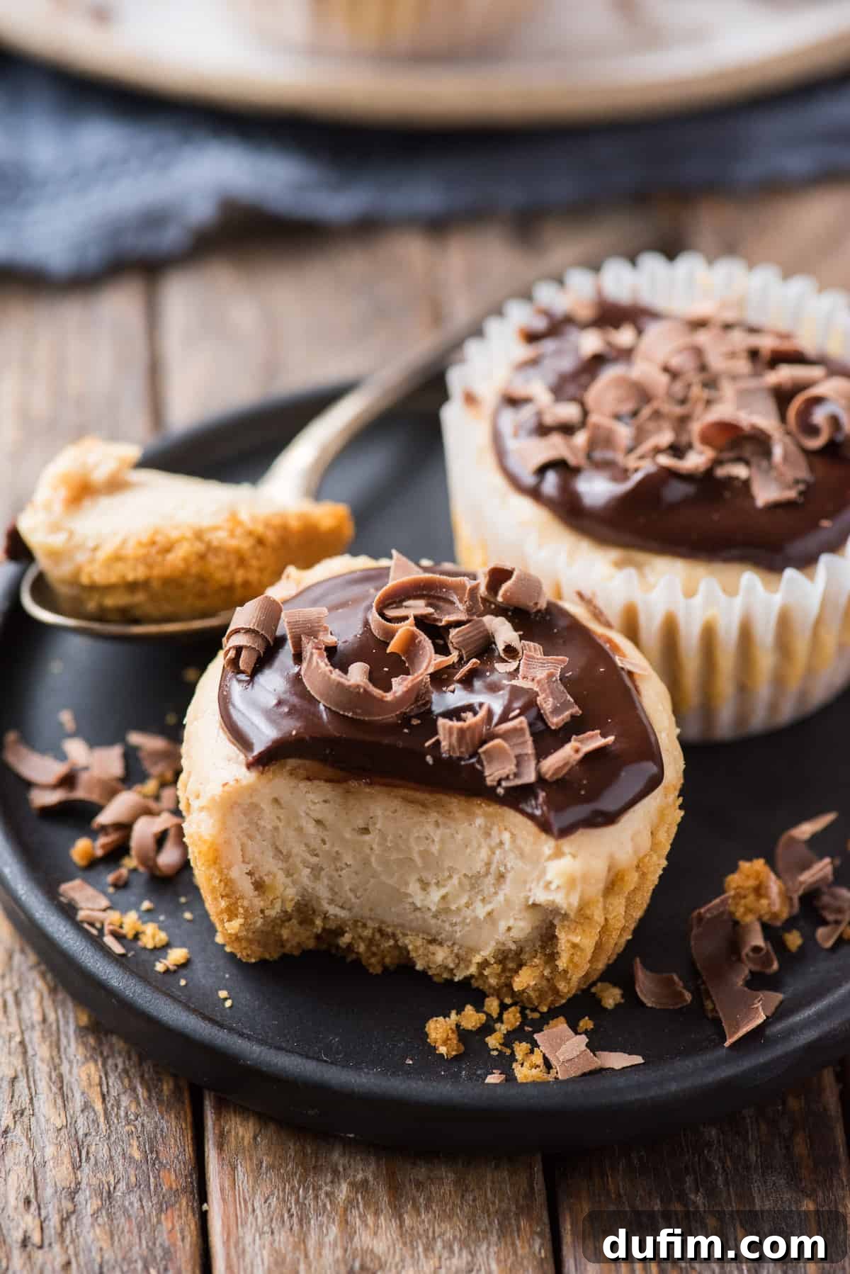 Peanut Butter Cheesecake Bites 2 Two perfectly portioned mini peanut butter cheesecakes adorned with a rich chocolate ganache topping, presented on a dark plate against a rustic wood background.