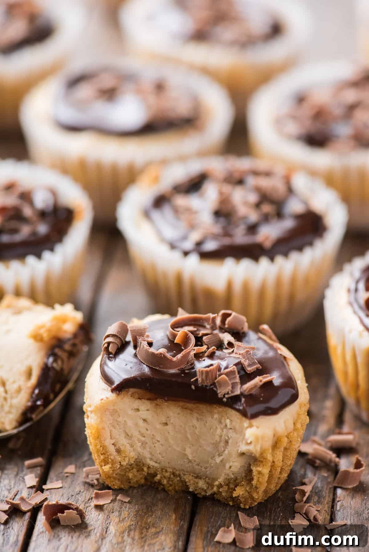 Peanut Butter Cheesecake Bites 6 An array of mini peanut butter cheesecakes with chocolate ganache topping, closely arranged on a wood background, with one cheesecake having a piece taken out.