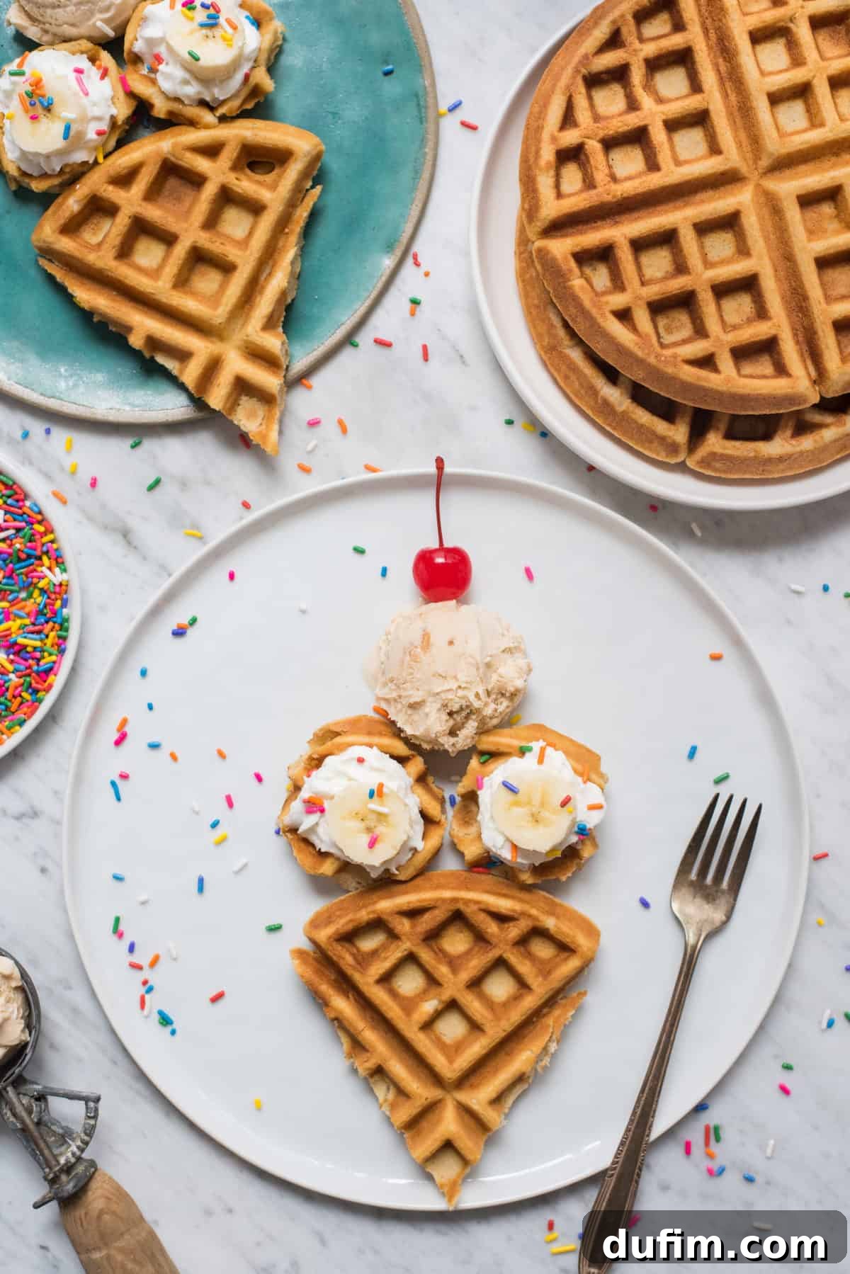 Waffles cut and arranged in the shape of an ice cream cone on a white plate, topped with banana slices, sprinkles, whipped cream, a scoop of ice cream, and a maraschino cherry. The plate is set against a clean white background.