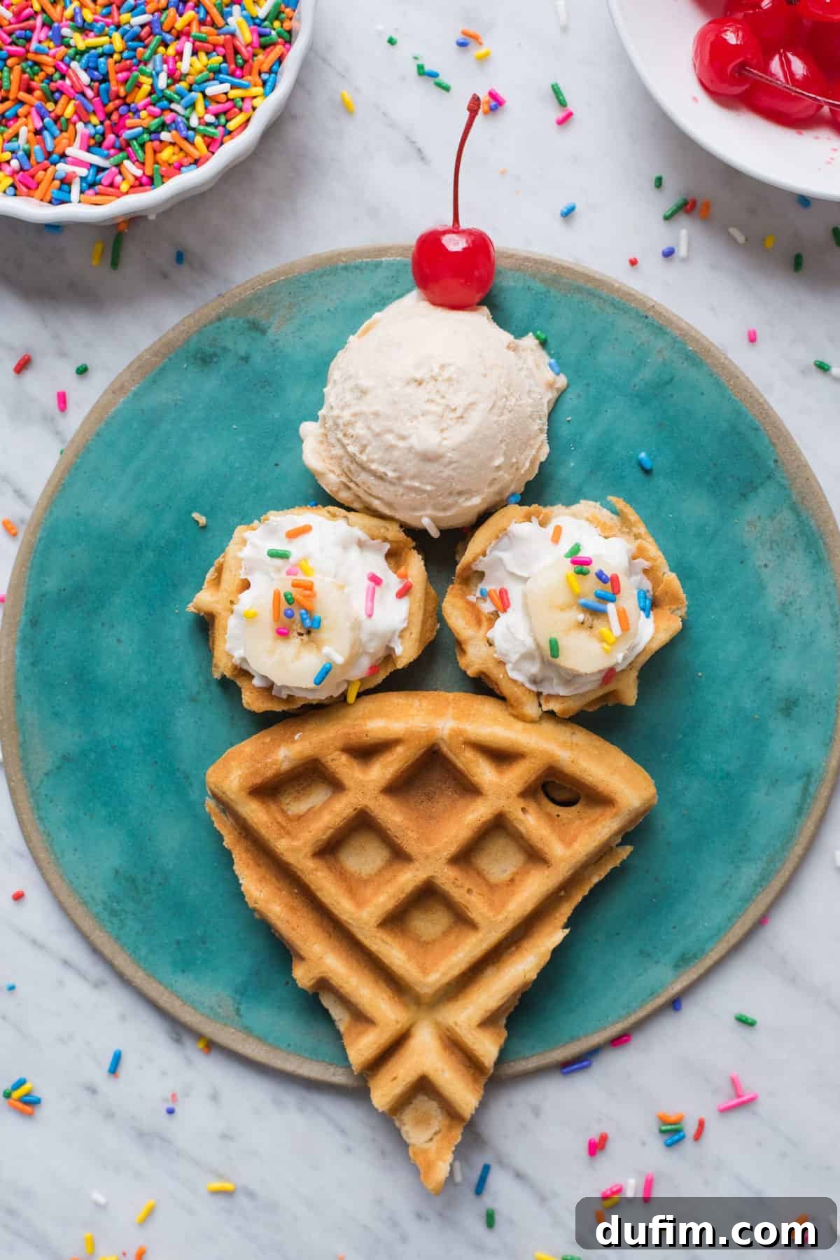 A vibrant teal plate holding two ice cream cone shaped waffles, each topped with a dollop of whipped cream, banana slices, and colorful sprinkles. A single scoop of ice cream sits beside them, adorned with a maraschino cherry. The plate is on a white background.