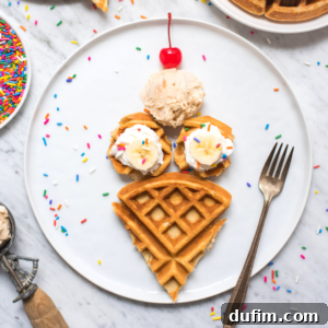 A plate with ice cream waffles arranged to look like a cone, topped with banana slices, sprinkles, whipped cream, a scoop of ice cream, and a cherry, with a fork on the side and sprinkles scattered around.