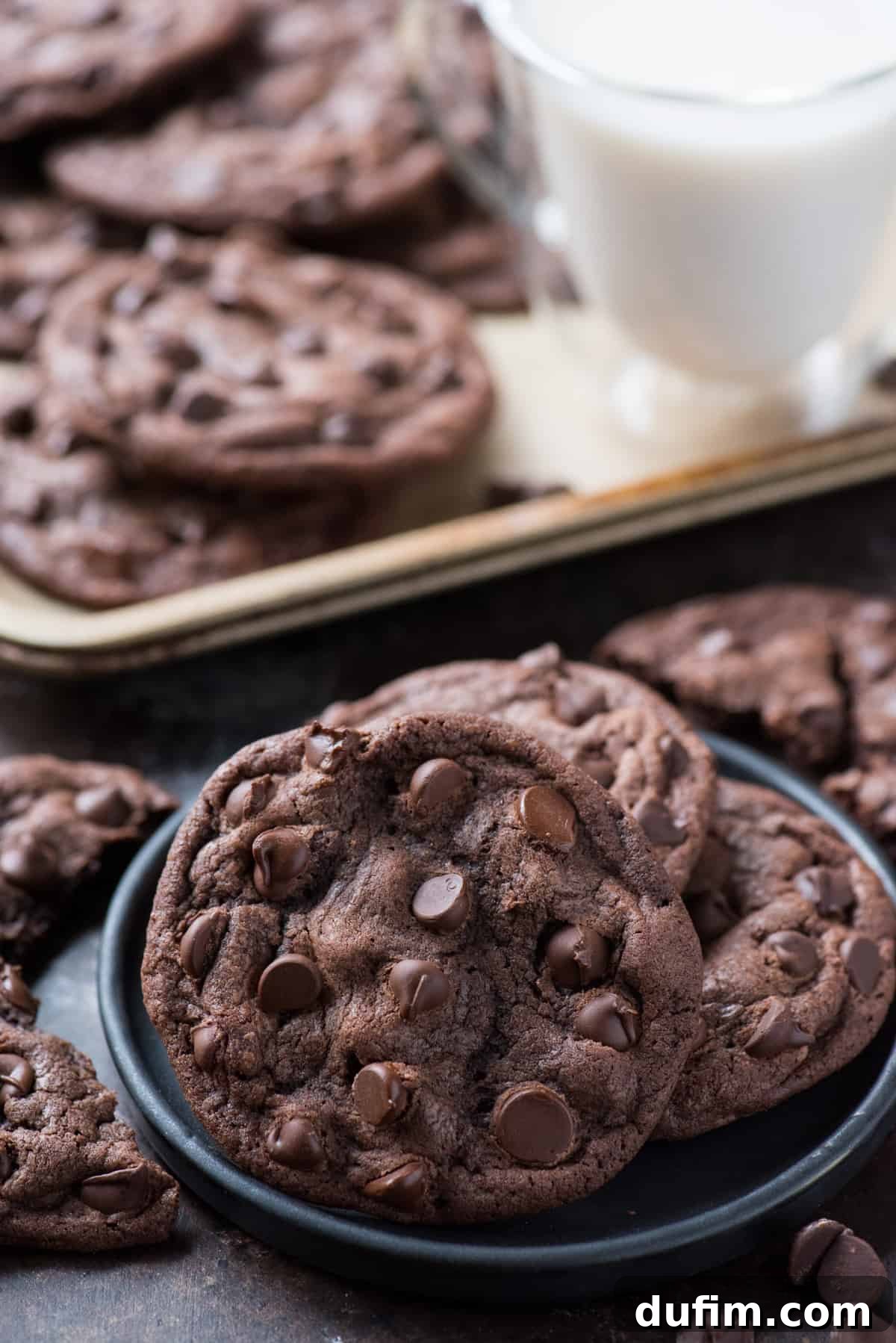Three perfectly baked double chocolate chip cookies resting on a dark navy plate, with a metal cookie sheet and a refreshing glass of milk blurred in the background.