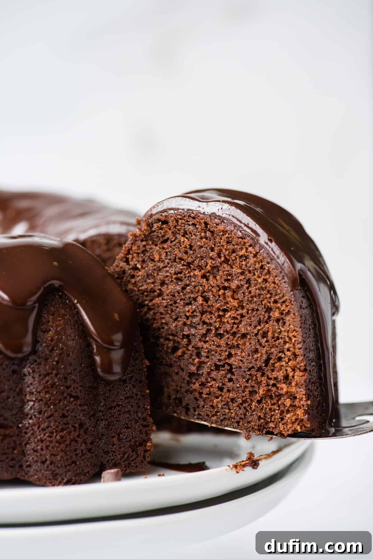 Decadent Chocolate Bundt with a Glossy Glaze 3 A delectable slice of chocolate bundt cake, generously covered in a glossy chocolate glaze, being carefully lifted from the main cake with a metal spatula against a bright white background.