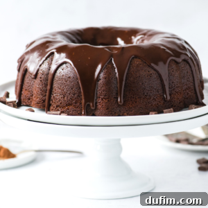 A rich chocolate bundt cake with glossy chocolate glaze dripping down the sides, beautifully displayed on a white cake stand.