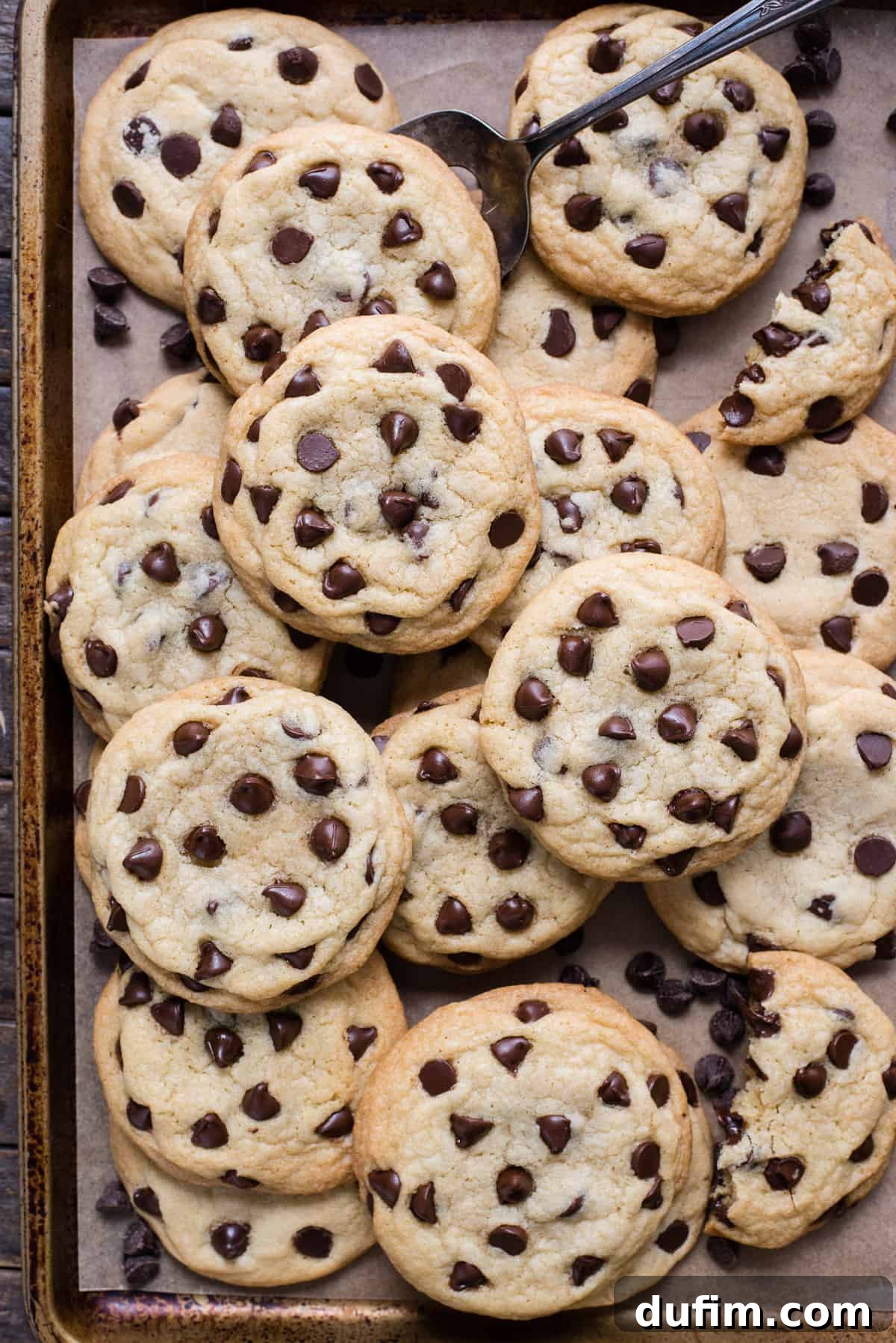 A pristine arrangement of golden-brown chocolate chip cookies, freshly baked and placed neatly on a metal baking sheet lined with brown parchment paper.