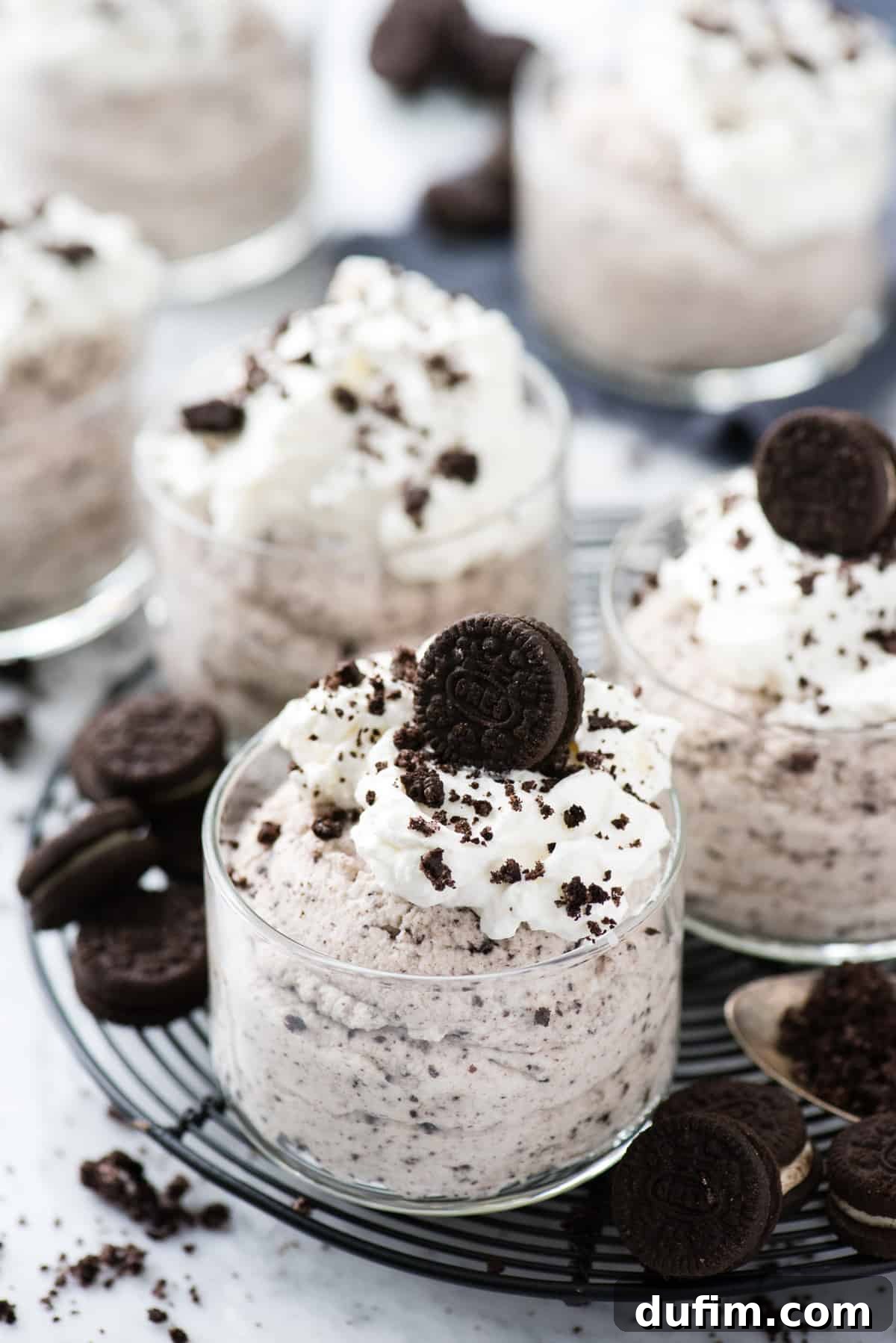 Creamy Oreo mousse in an elegant glass dessert bowl, topped with a generous swirl of whipped cream and sprinkled with crushed Oreo pieces, resting on a stylish black circular rack on a clean white background. This image perfectly captures the luxurious texture and delicious appeal of a homemade no-bake dessert.