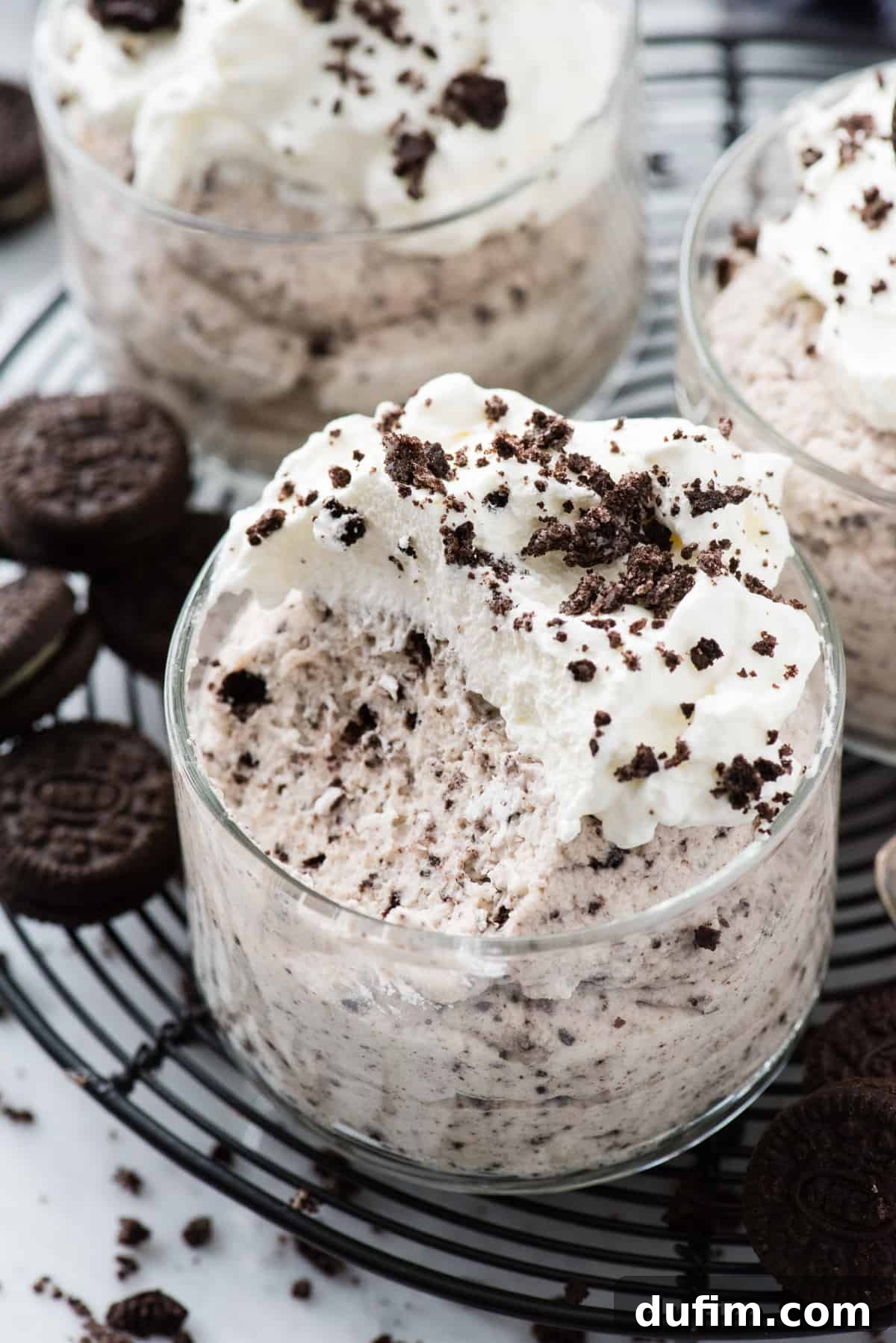 A glass bowl filled with rich, dark Oreo mousse, with a spoonful already removed, revealing the fluffy texture. It's beautifully topped with a swirl of homemade whipped cream and scattered Oreo crumbs, presented on a black wire rack against a soft white background. This image highlights the irresistible indulgence of this simple dessert.