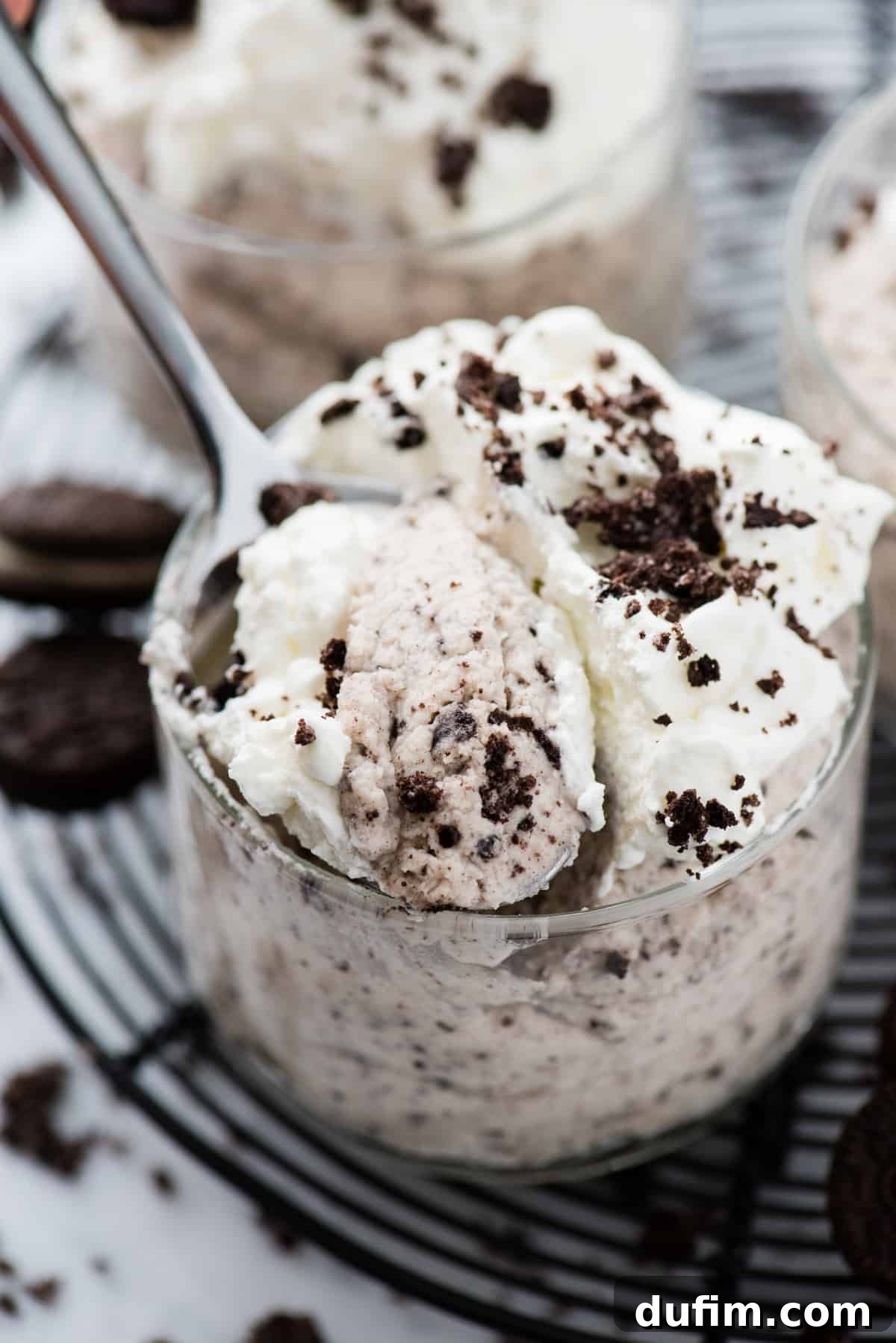 A glass bowl of fluffy Oreo mousse, with a spoon gently resting inside, suggesting a taste has just been taken. The mousse is generously topped with creamy whipped cream and crushed Oreo crumbs, presented on a black wire rack against a soft white background. This photo invites you to indulge in its rich and airy texture.