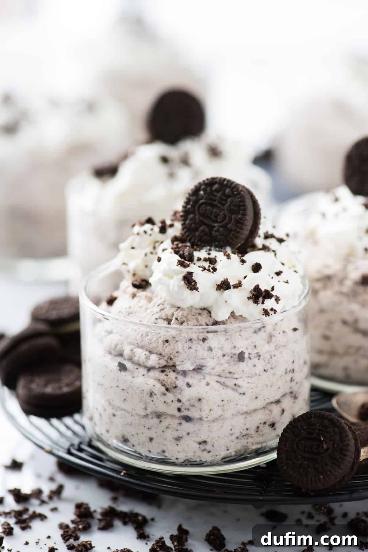 Five glass bowls filled with creamy Oreo mousse, each exquisitely topped with a swirl of fresh whipped cream and a sprinkle of crushed Oreo pieces, all arranged on a sophisticated black circular wire rack against a pristine white background. This inviting display highlights the dessert's elegance and its suitability for serving to guests.