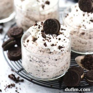 Glass cups filled with rich oreo mousse, topped with whipped cream, crushed cookie crumbs, and a mini chocolate sandwich cookie, displayed on a wire rack. Other cookies and crumbs are scattered around.