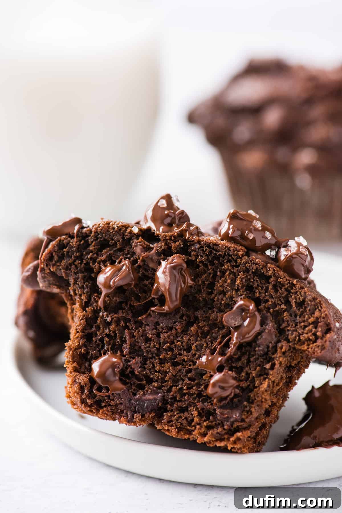 Decadent Chocolate Muffins 8 A double chocolate chip muffin, expertly sliced in half, reveals a perfectly moist texture brimming with melted chocolate chips, presented on a white plate against a clean white background.