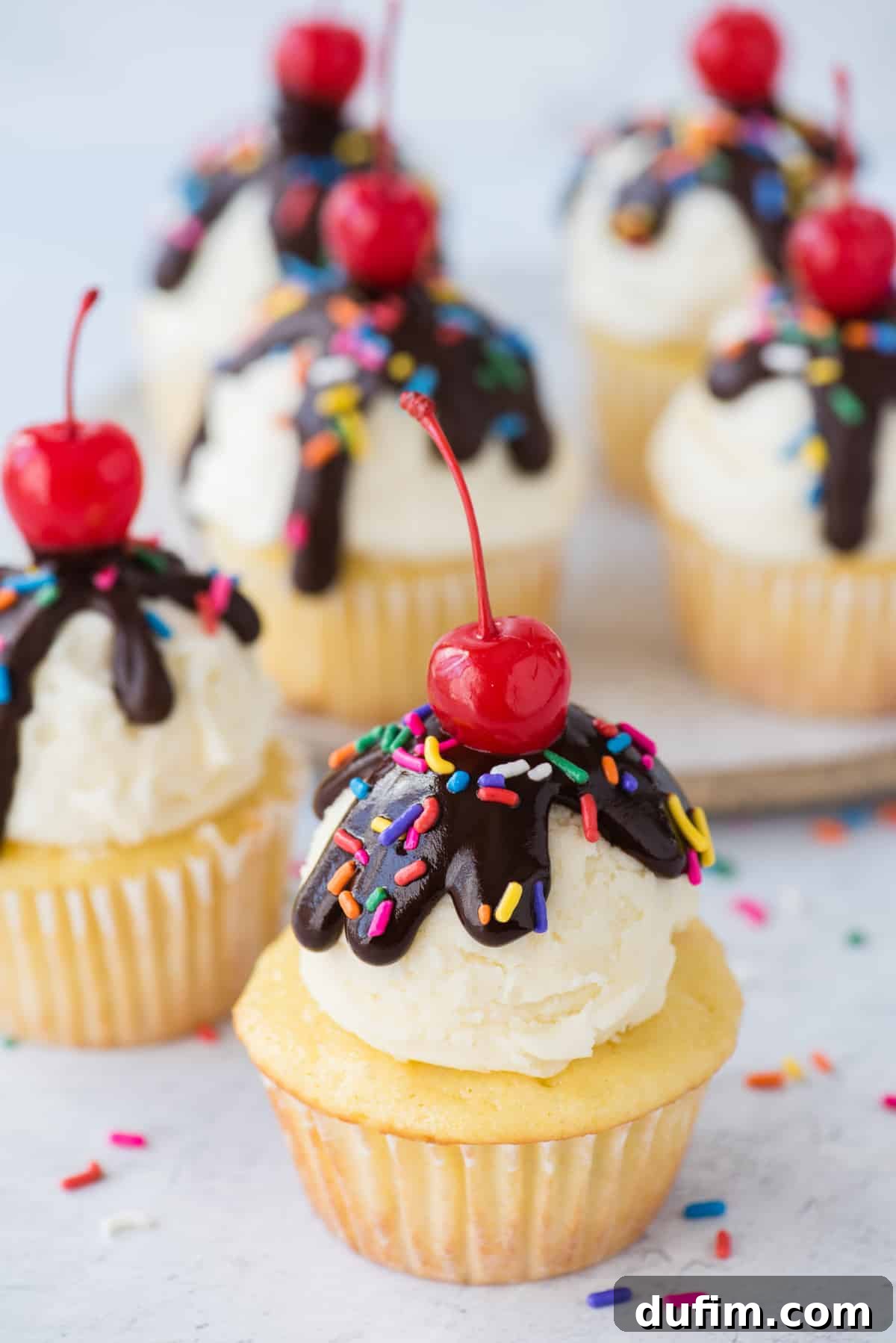 A beautifully decorated birthday cupcake with a generous scoop of smooth vanilla frosting, glistening chocolate ganache drizzle, vibrant sprinkles, and a classic maraschino cherry on top, set against a clean white background.