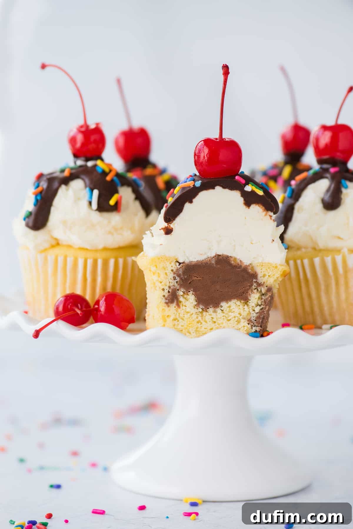 An ice cream cupcake cut in half, revealing a luscious chocolate ice cream center, displayed alongside whole ice cream cupcakes on a white cake stand against a white background.