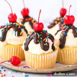 Delicious vanilla ice cream cupcakes topped with white frosting, a rich chocolate drizzle, vibrant rainbow sprinkles, and a classic red cherry. They are artfully arranged on a plate with extra cherries and sprinkles scattered around, inviting you to indulge.