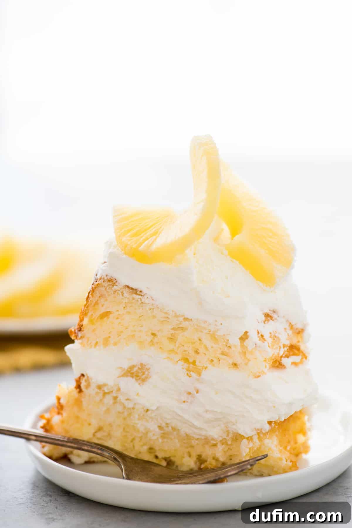 A slice of fluffy pineapple angel food cake layered with whipped cream and pineapple pieces on a white plate, set against a pristine white background.