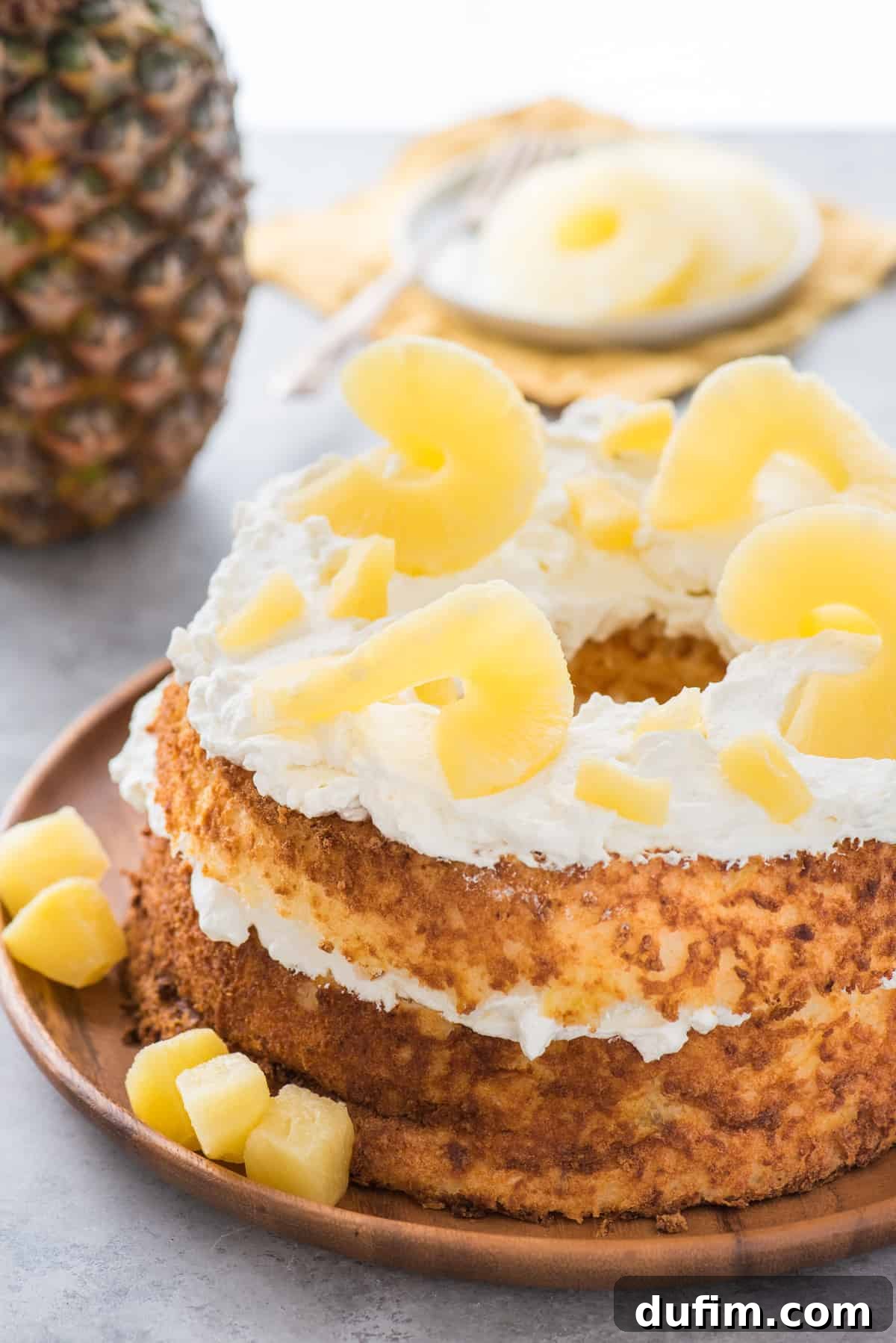 A beautifully decorated whole angel food cake topped with swirls of whipped cream and fresh pineapple slices, served on a rustic wooden tray, ready for slicing.