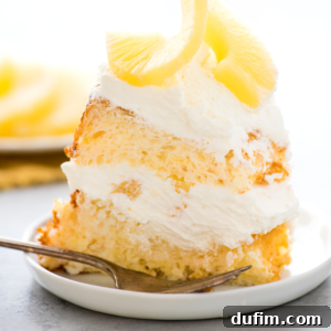 A slice of fluffy pineapple angel food cake layered with whipped cream and topped with pineapple slices sits on a white plate with a fork beside it.