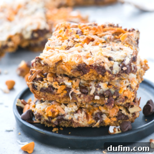 A close-up of three 7 layer bars stacked on a black plate, topped with shredded coconut, chocolate chips, and butterscotch chips. Crumbs and extra chips are scattered around on a light surface.