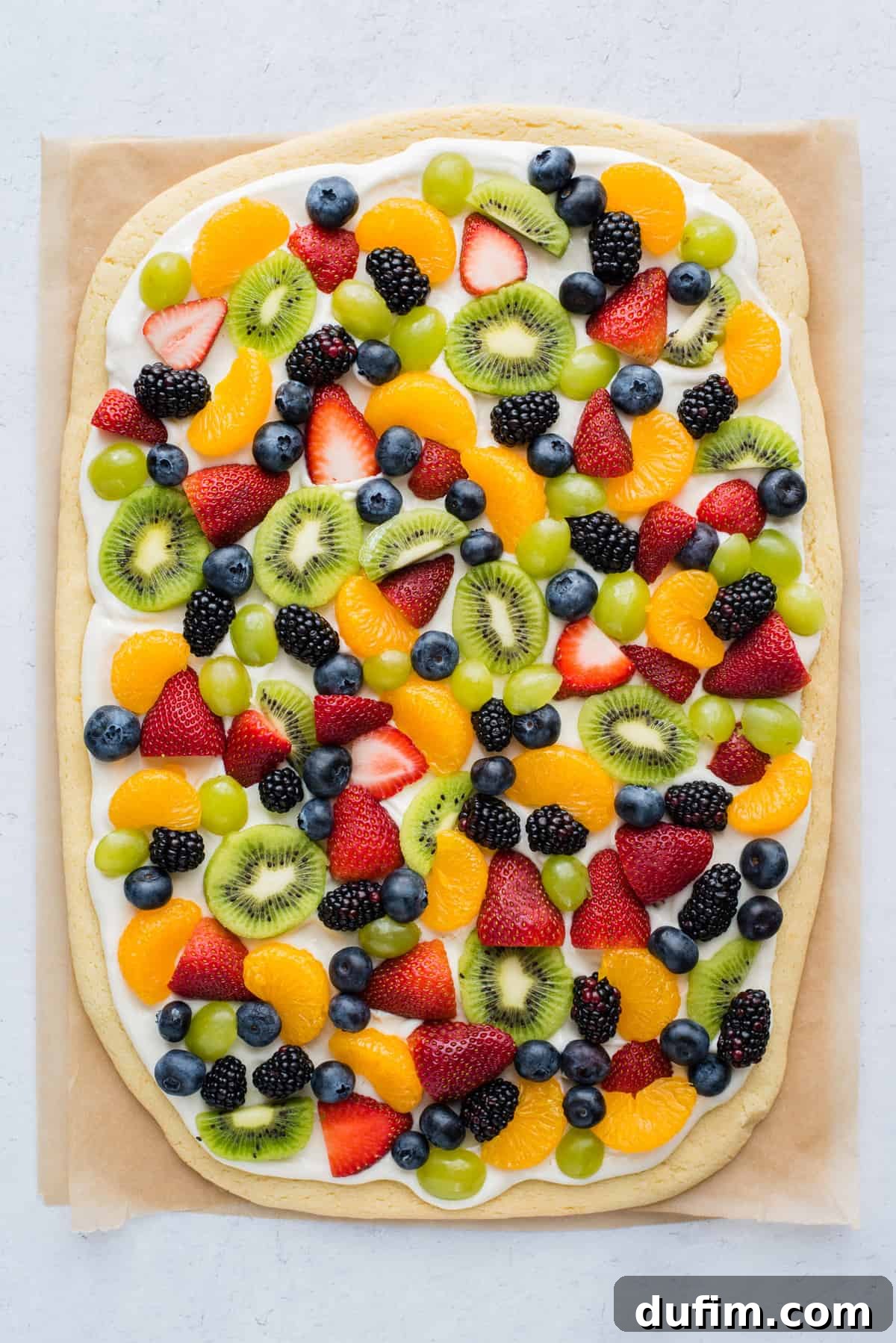 A vibrant whole fruit pizza adorned with fresh strawberries, blueberries, mandarin oranges, kiwi slices, and green grapes, resting on brown parchment paper.