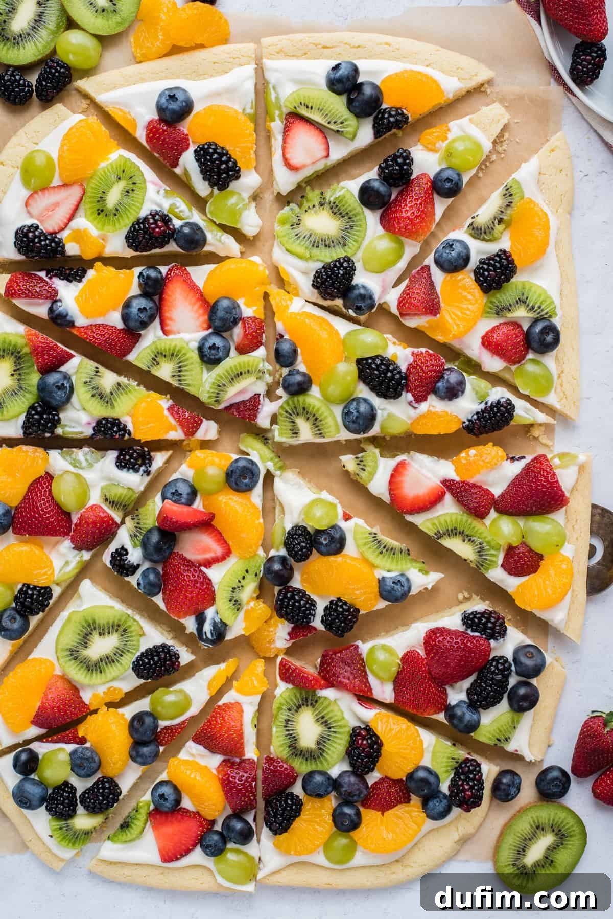 A whole fruit pizza, perfectly cut into triangle slices, topped with a colorful array of strawberries, blueberries, mandarin oranges, kiwi, and grapes, displayed on brown parchment paper.