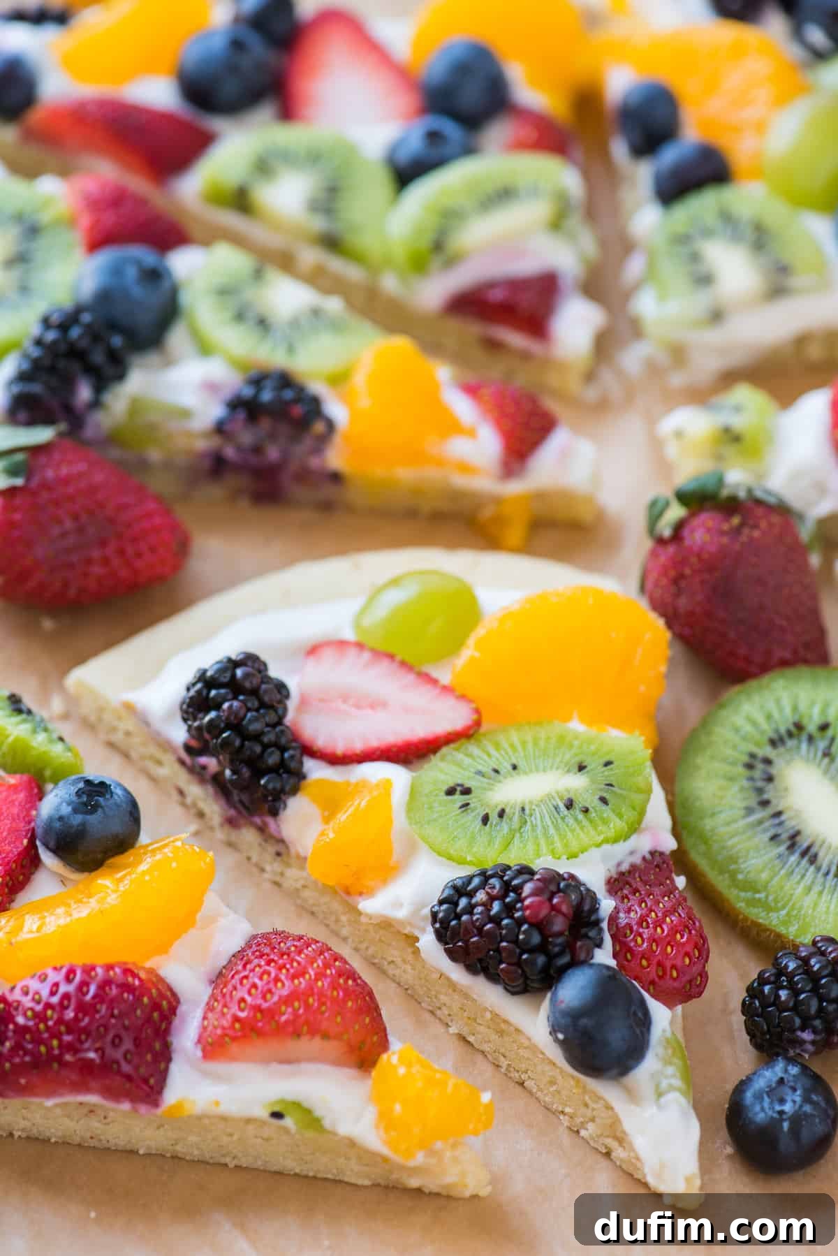 A delicious single slice of fruit pizza topped with strawberries, blueberries, mandarin oranges, kiwi, and grapes, presented on brown parchment paper.