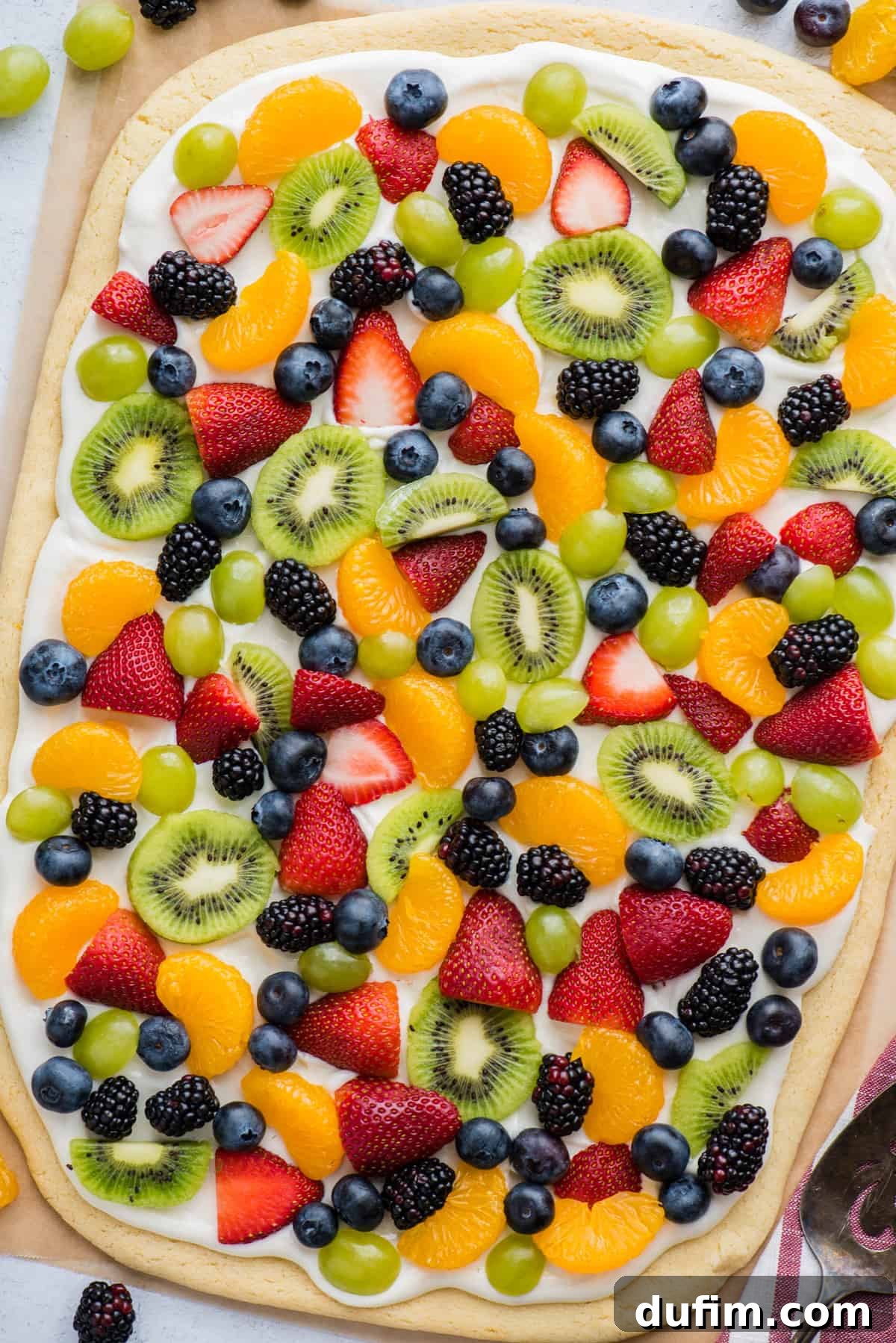 A beautifully arranged whole fruit pizza featuring strawberries, blueberries, mandarin oranges, kiwi, and grapes, all on a sugar cookie crust and brown parchment paper background.