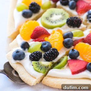 A close-up of a delicious fruit pizza slice topped with cream cheese frosting, blueberries, blackberries, kiwi, strawberries, green grapes, and orange segments on a soft sugar cookie crust.