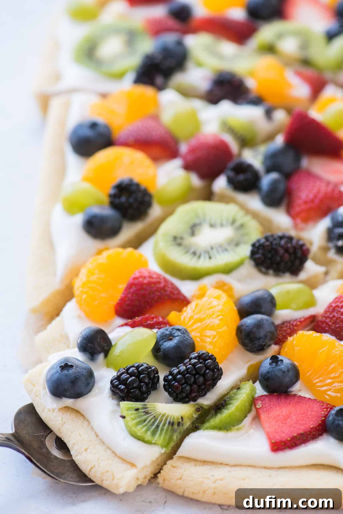A perfect slice of fruit pizza, showcasing its creamy frosting and vibrant fruit toppings, being lifted from the pan with a metal spatula.