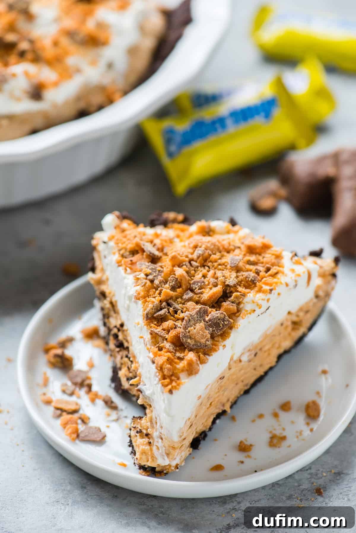 Butterfinger Crunch Pie 2 A perfectly sliced piece of Butterfinger pie on a white plate, with the rest of the pie in a pan blurred in the background, showcasing its rich layers and tempting texture.