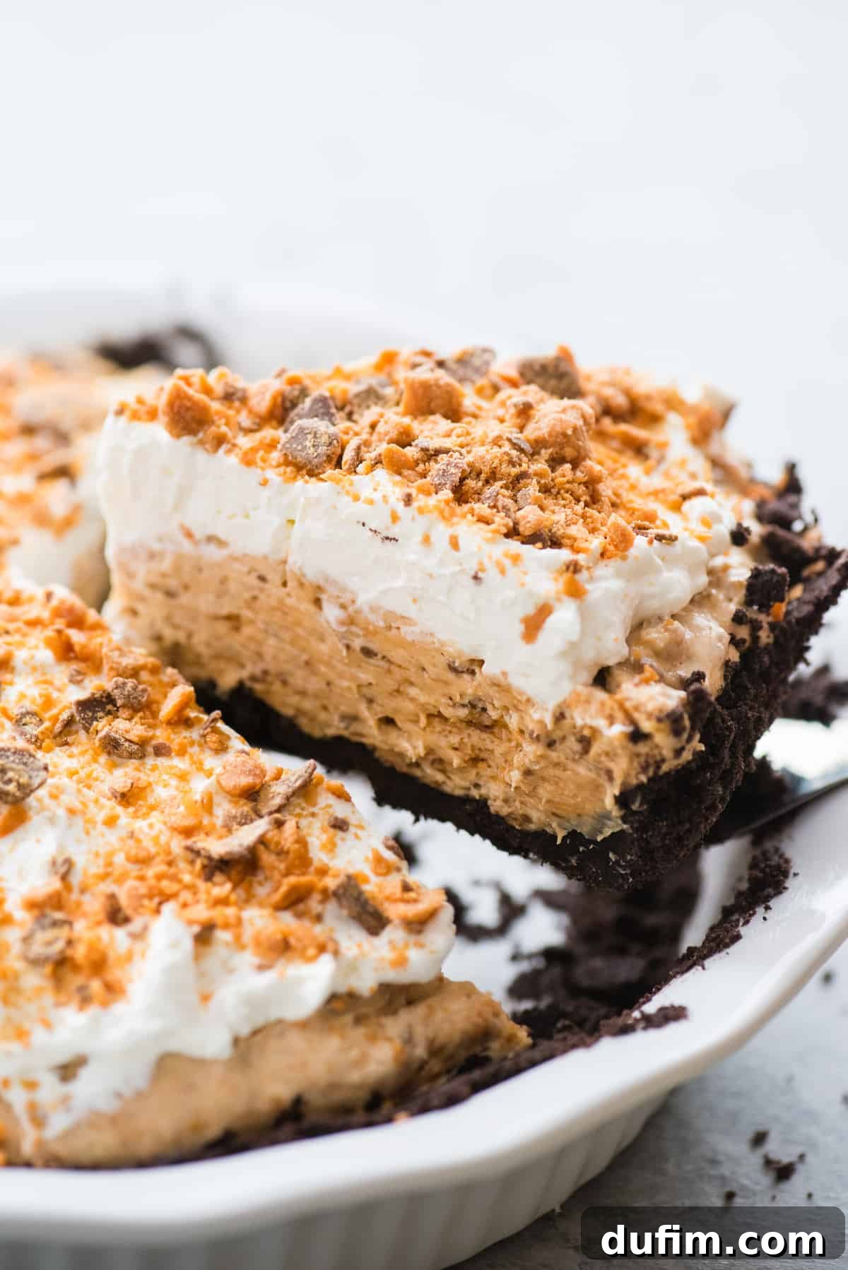 Butterfinger Crunch Pie 5 A slice of creamy Butterfinger pie being carefully lifted from a white pie pan with a spatula, showing its distinct layers and tempting texture.