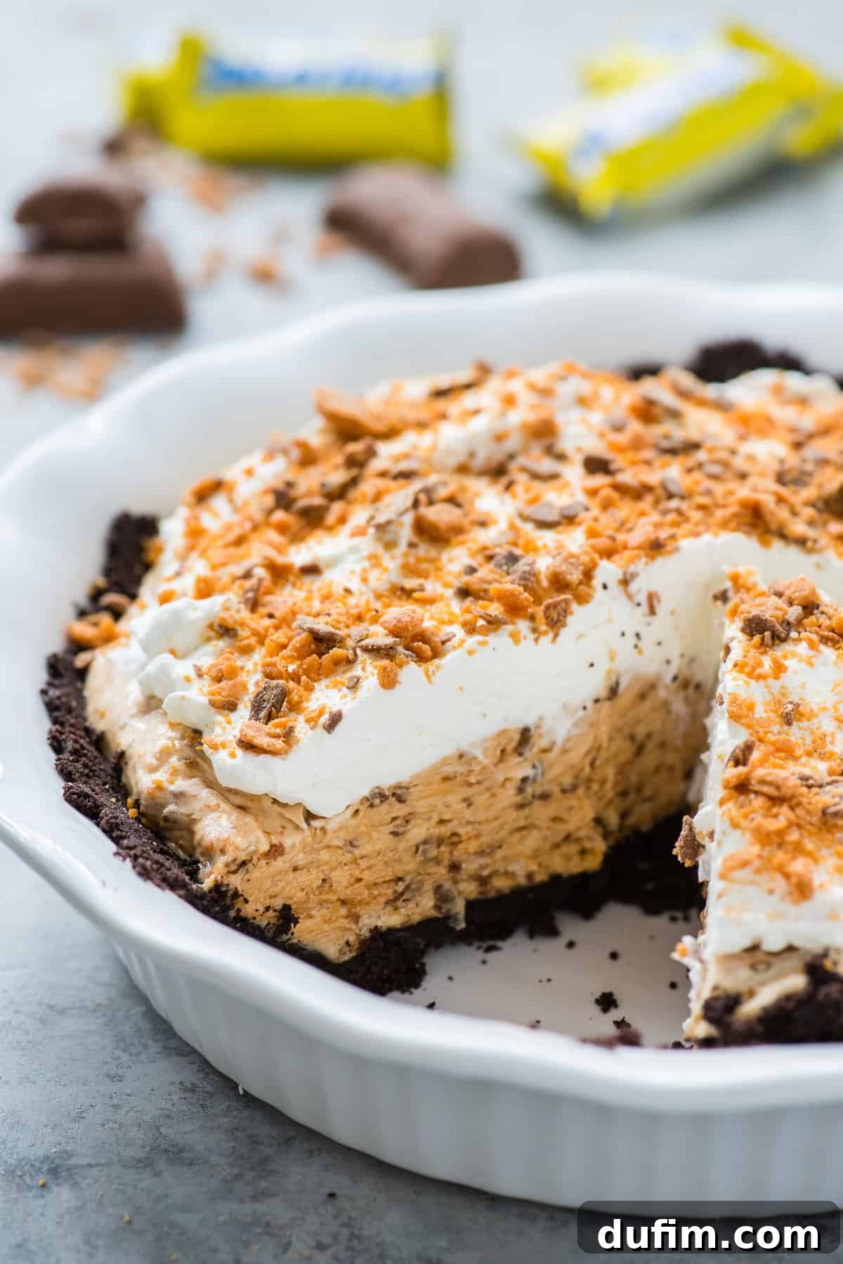 Butterfinger Crunch Pie 7 A whole Butterfinger pie in a white pie pan with one slice already removed, showcasing its layers, against a subtle gray background.