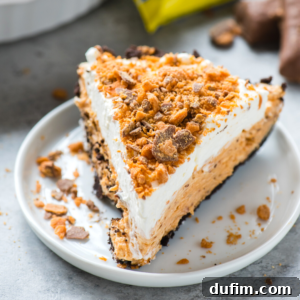 A slice of creamy Butterfinger pie with a crumbly chocolate crust, topped with whipped cream and crushed candy pieces, sits on a white plate. Some crumbs are scattered around the plate.