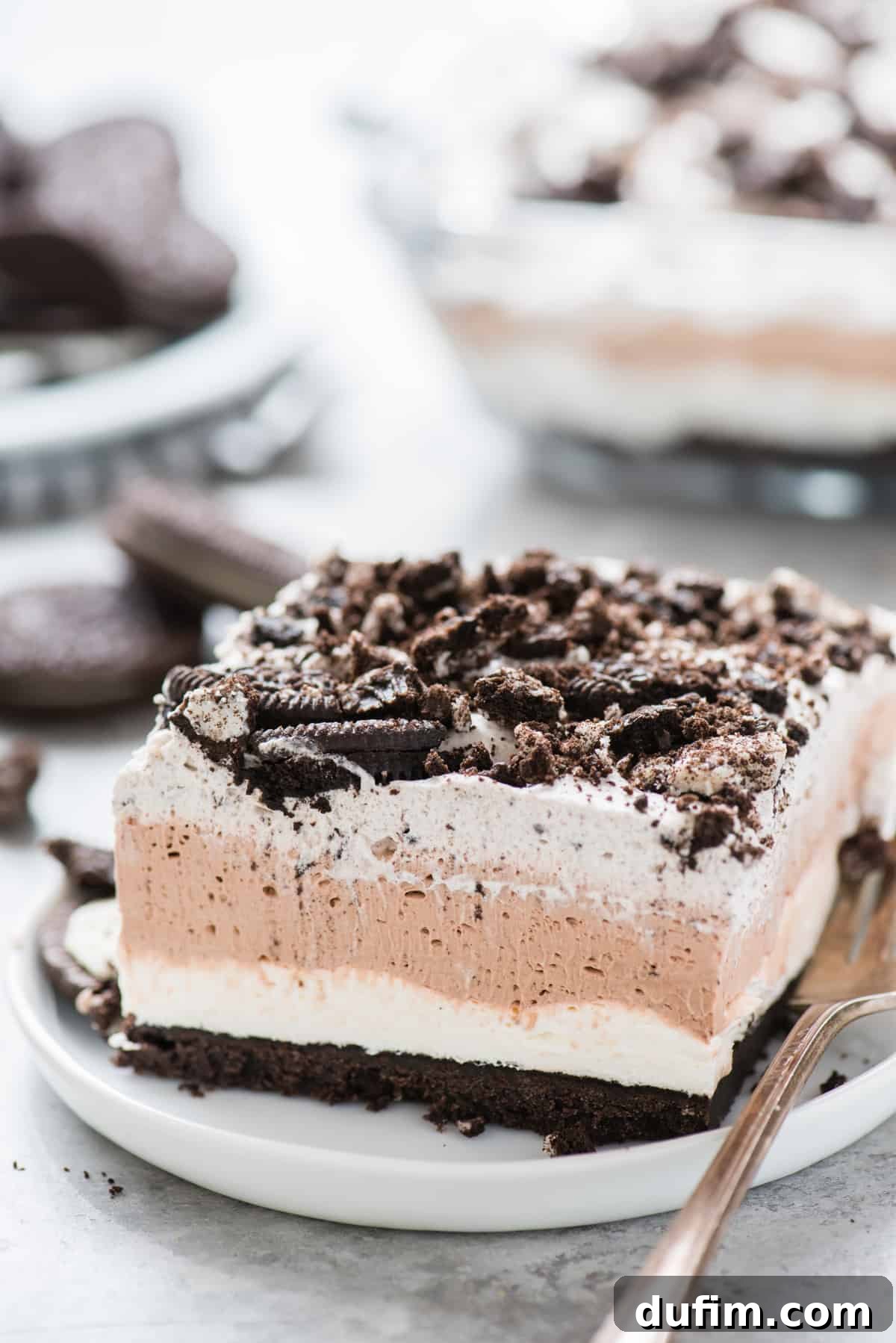 A perfectly cut slice of no-bake Oreo layer dessert, showcasing a rich dark Oreo crust, a creamy white filling, a thick chocolate mousse layer, and a light whipped topping, garnished with Oreo crumbles, all resting on a pristine white plate against a minimalist white background. This image highlights the dessert's clean layers and enticing texture.