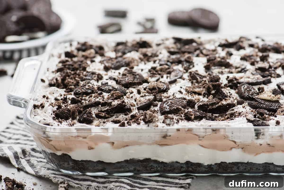 A pristine 9x13 inch glass baking dish filled with the layered no-bake Oreo dessert, showing the various dark and light creamy layers, ready to be chilled and served. A blue striped napkin is placed artfully underneath the pan, suggesting a summer or casual setting.