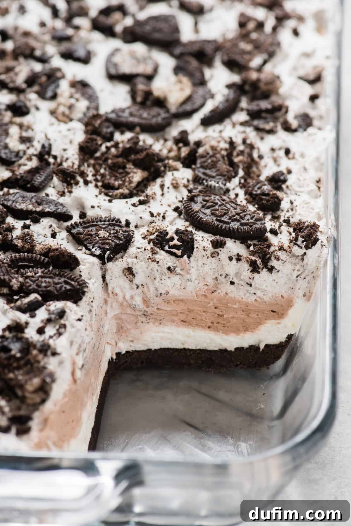 A pristine, perfectly cut slice of layered no-bake Oreo dessert has been removed from a clear glass 9x13 inch pan, showcasing the distinct, firm layers of dark Oreo crust, light cream cheese, rich chocolate mousse, and fluffy Oreo whipped cream. The clean lines emphasize the dessert's excellent structural integrity.