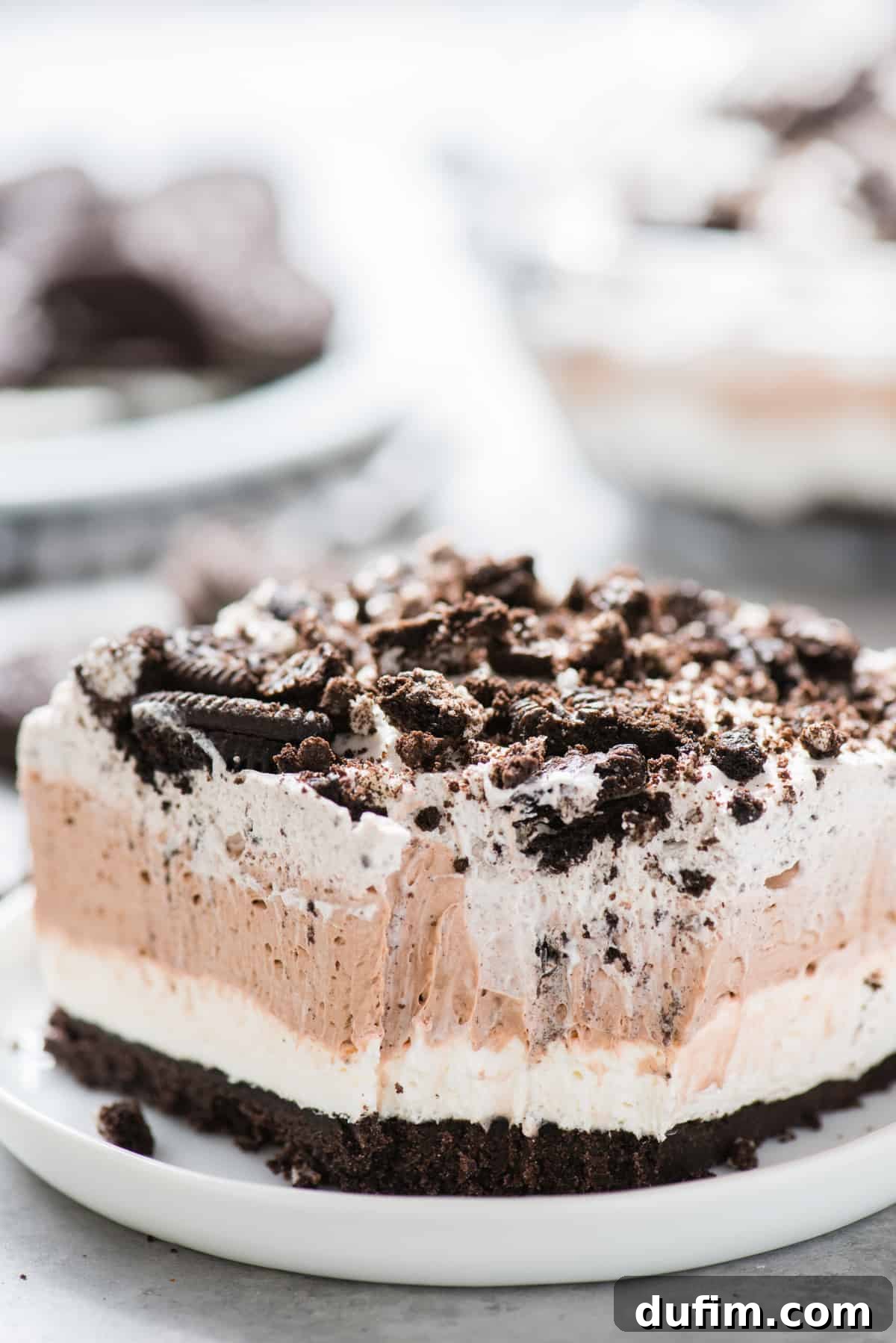 A tempting slice of no-bake Oreo layer dessert, taken with a small bite removed, sits on a clean white plate. The visible layers, from the dark Oreo crust to the fluffy whipped cream, indicate its rich texture and deliciousness. The white background emphasizes the dessert as the focal point.