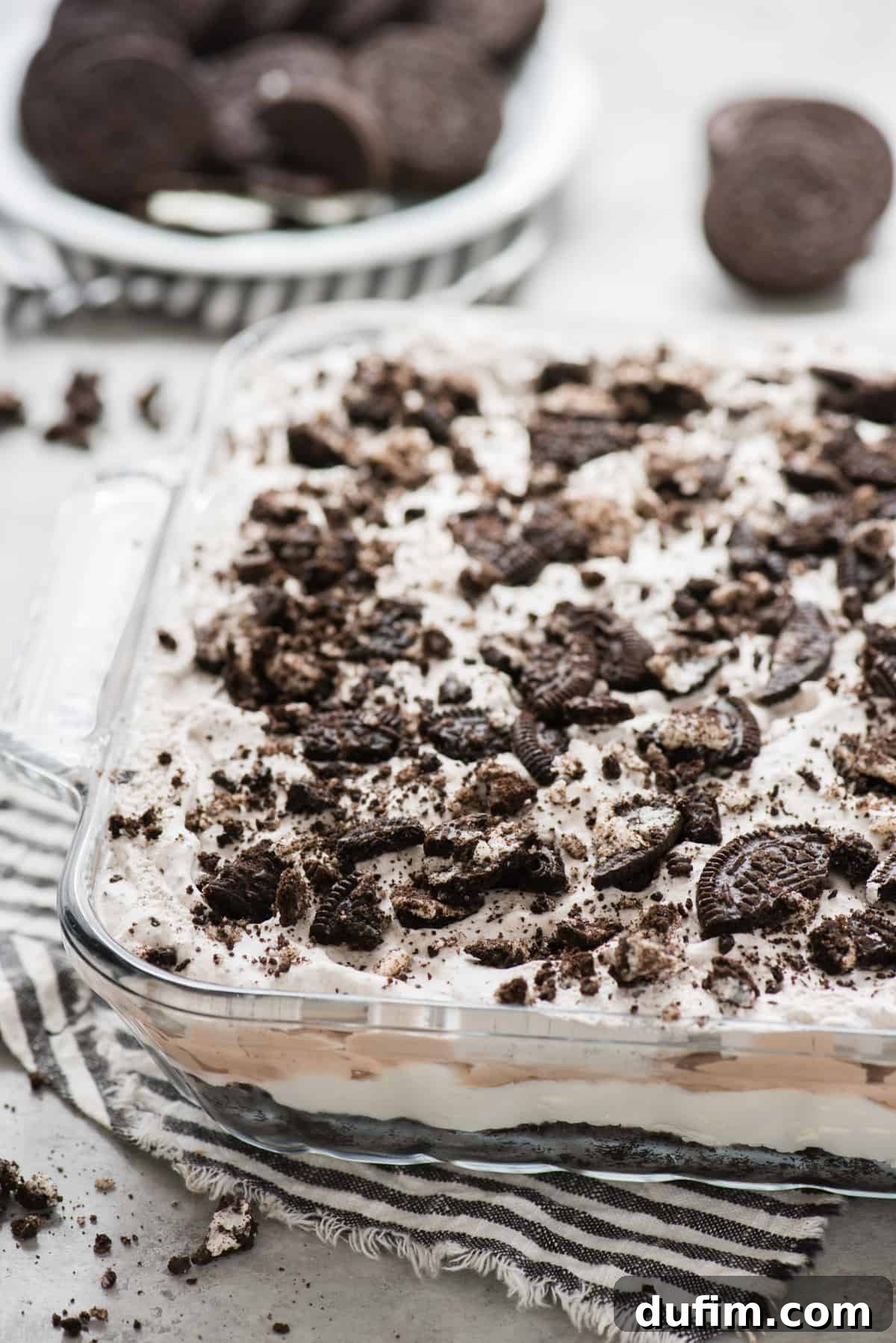 The fully assembled No-Bake Oreo Dessert, presented in a clear glass 9x13 inch pan, awaiting its chilling period. The distinct layers are visible and topped with a generous scattering of crushed Oreos. A blue striped napkin accentuates the pan's presentation.
