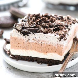 A slice of layered Oreo dessert on a white plate, showcasing a dark Oreo crust, creamy white layer, chocolate mousse, and whipped cream topping with cookie crumbles. A fork rests beside it, indicating readiness to eat.