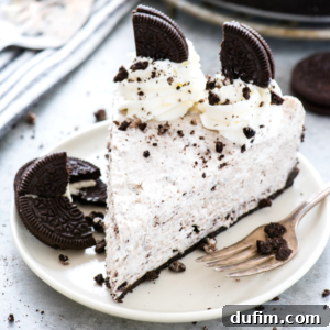 A slice of no-bake Oreo cheesecake topped with whipped cream, cookie crumbles, and two cookie halves, served on a white plate with a fork and extra cookie pieces on the side.