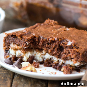 A close-up of an earthquake cake-inspired chocolate brownie with a creamy white filling and scattered chocolate chips, served on a white plate atop a rustic wooden surface.