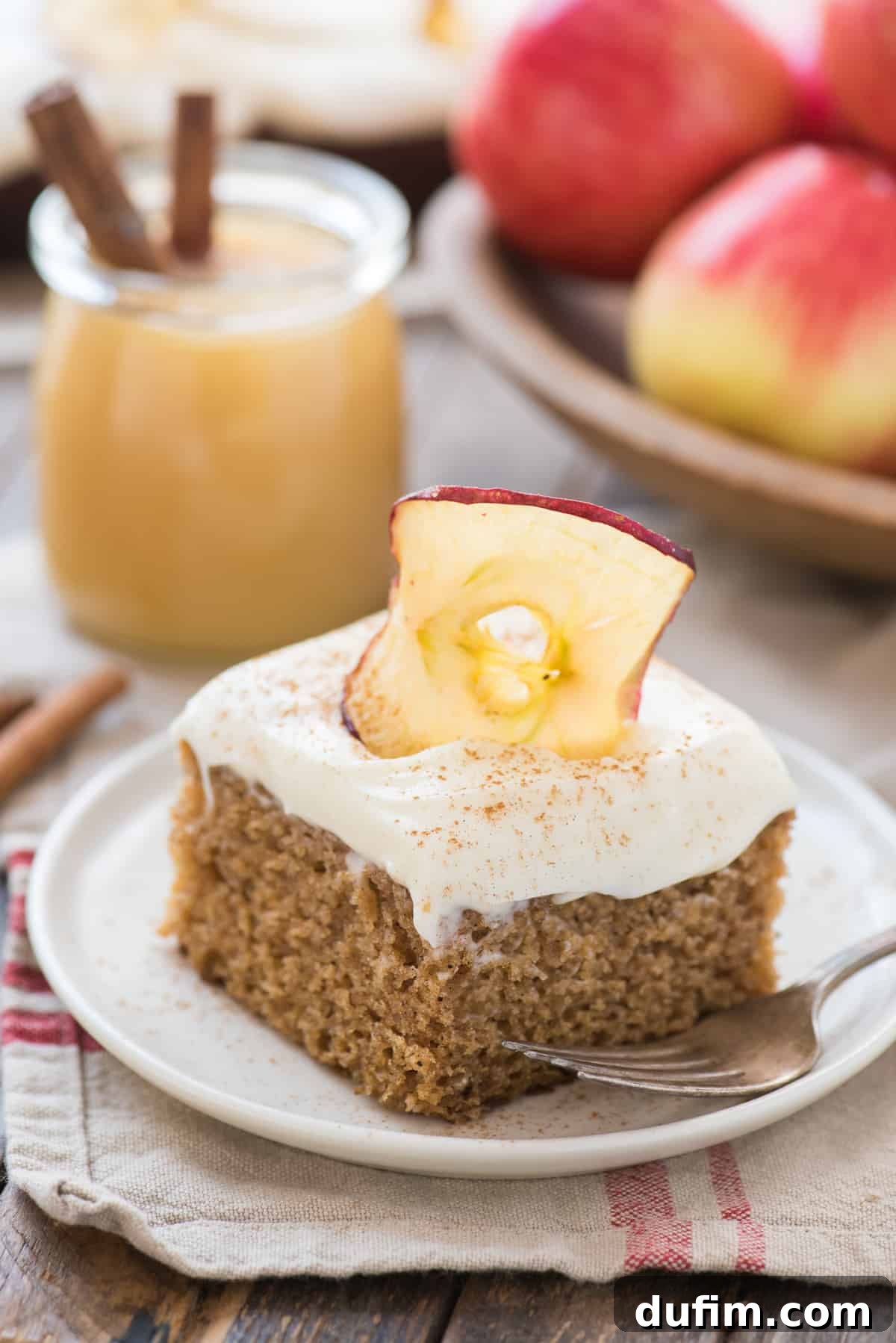 Warm Applesauce Spice Cake 2 A delectable slice of applesauce cake topped with creamy white frosting, served on a white plate with fresh red apples blurred in the background, signaling the essence of fall.
