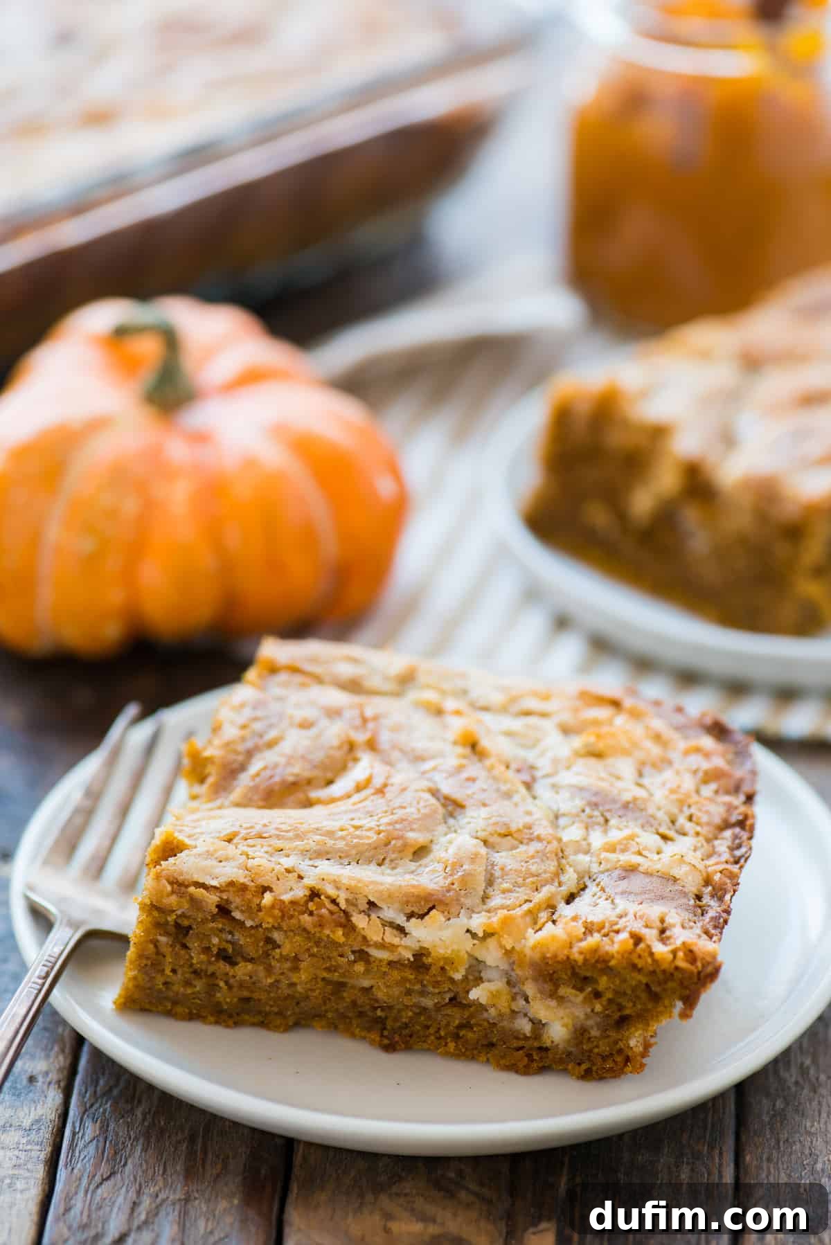 Spiced Pumpkin Earthquake Cake 5 A close-up of a delectable slice of pumpkin earthquake cake on a white plate, with a rustic pumpkin decoration blurred in the background, emphasizing the cake's autumnal appeal.