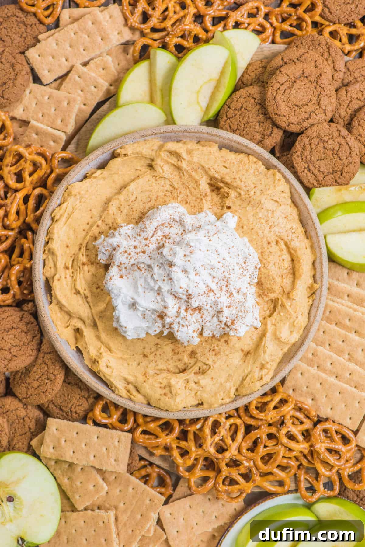 A bowl of creamy pumpkin dip topped with whipped cream, surrounded by delicious dippers like pretzels, gingersnaps, graham crackers, and crisp green apple slices, set against a warm, inviting backdrop.