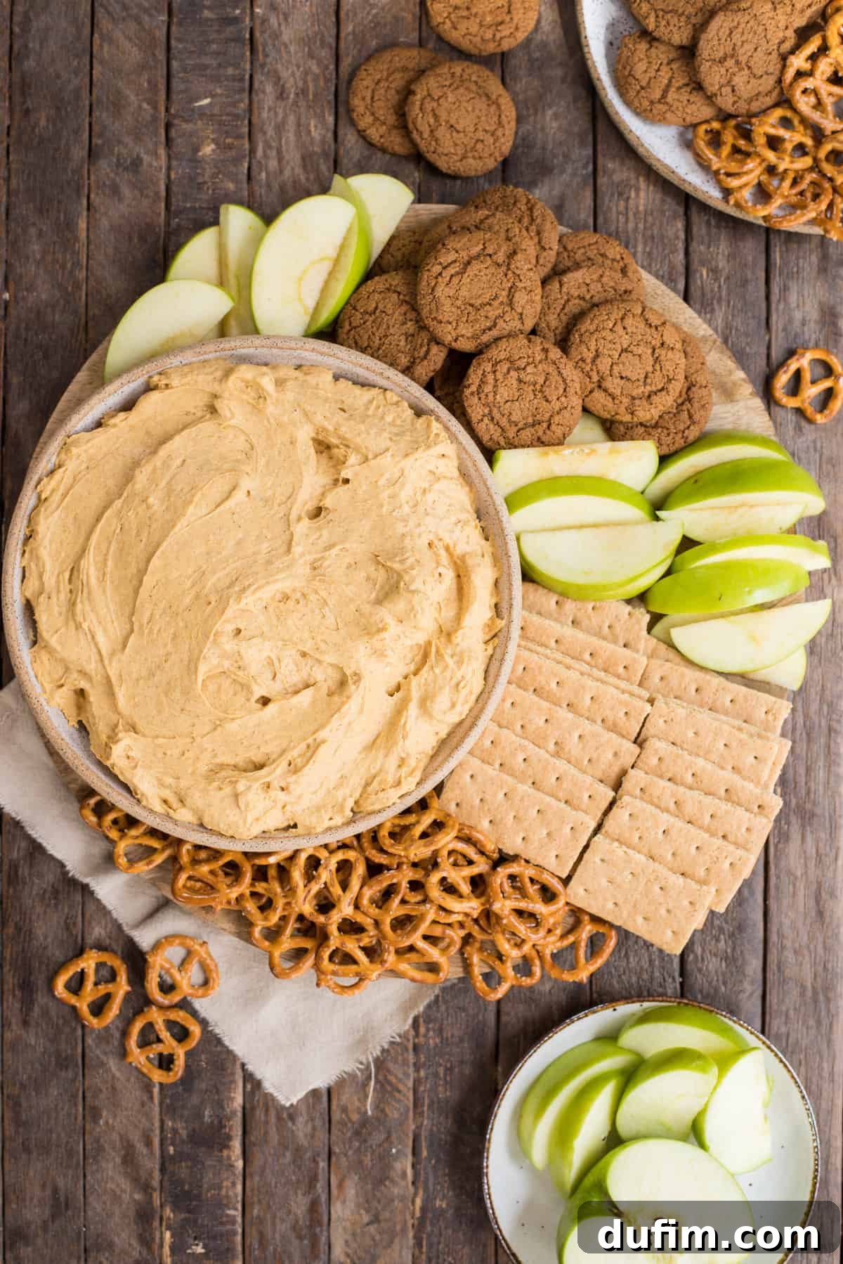 A beautifully presented bowl of pumpkin dip, garnished with a dollop of whipped cream and a sprinkle of cinnamon, surrounded by an array of dippers including pretzels, gingersnaps, graham crackers, and fresh green apple slices, all resting on a rustic wooden surface.