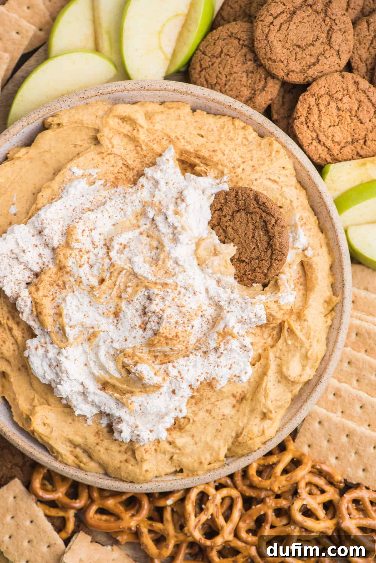 An overhead shot of the alluring pumpkin dip, artfully garnished with a swirl of whipped cream, sitting proudly amidst a delicious spread of crunchy pretzels, spiced gingersnaps, classic graham crackers, and crisp green apple slices.